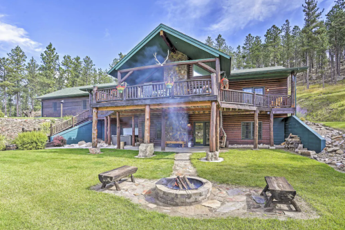 A large log cabin with a spacious porch, surrounded by trees and a fire pit in the foreground on a sunny day.