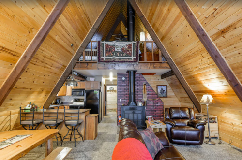Cozy A-frame cabin interior with wooden walls, a stone fireplace, kitchen bar, and comfortable seating area.