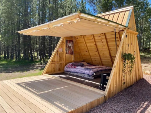 A cozy wooden A-frame cabin with an open front, featuring a bed and surrounded by trees.