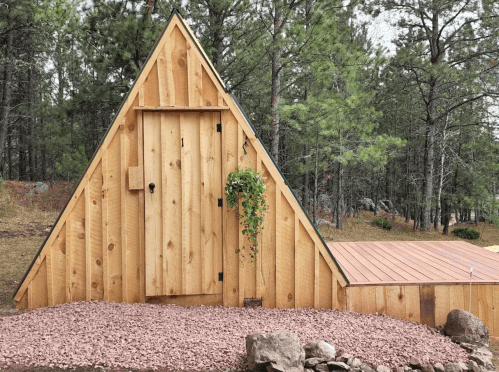A wooden A-frame cabin with a door and a hanging plant, surrounded by gravel and trees in a natural setting.