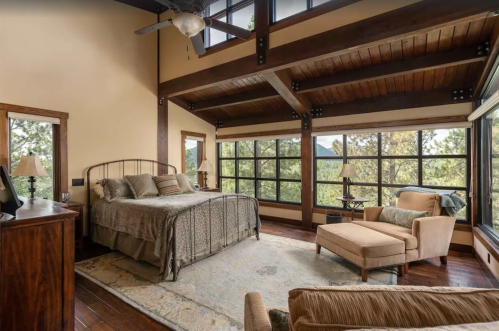 Cozy bedroom with large windows, wooden beams, a bed, and a lounge chair, offering a scenic view of nature.