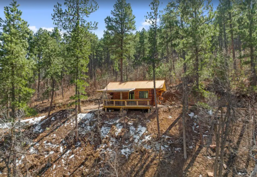 A cozy log cabin nestled among tall pine trees on a hillside, with a small porch and patches of snow on the ground.