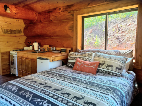 Cozy cabin interior with a bed, wooden kitchen area, and large window showcasing nature outside.