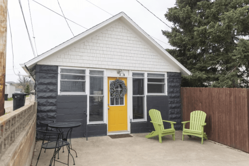 A small house with a yellow door, black and white exterior, and two green chairs on a concrete patio.
