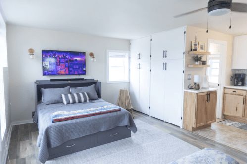A cozy bedroom featuring a bed, wall-mounted TV, and kitchenette with wooden cabinets and a light color scheme.