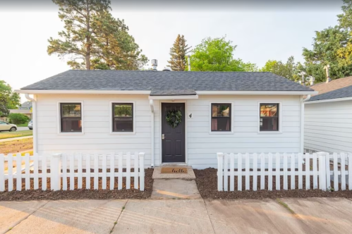 A small white house with a gray door, surrounded by a white picket fence and greenery in the background.