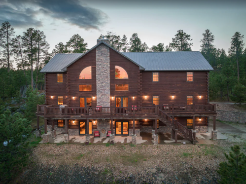 A large wooden cabin with a stone facade, surrounded by trees, featuring multiple decks and large windows at dusk.