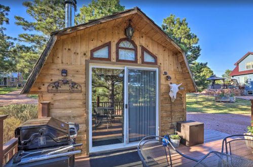 A rustic wooden cabin with a sliding glass door, surrounded by trees and outdoor seating on a sunny day.