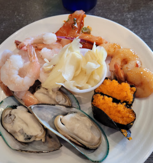 A plate of assorted seafood including shrimp, oysters, and sushi topped with orange roe, accompanied by pickled ginger.