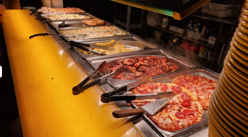 A buffet table with various pizzas, pasta, and dishes in metal trays under warm lighting.