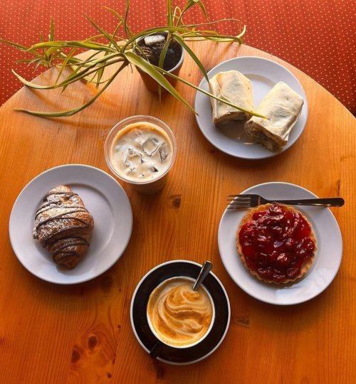 A wooden table with two plates of pastries, a coffee cup, and an iced drink, alongside a small potted plant.