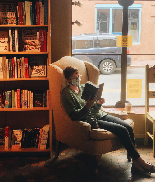 A person sits in a cozy chair reading a book in a bookstore, with shelves of books and a window view behind them.