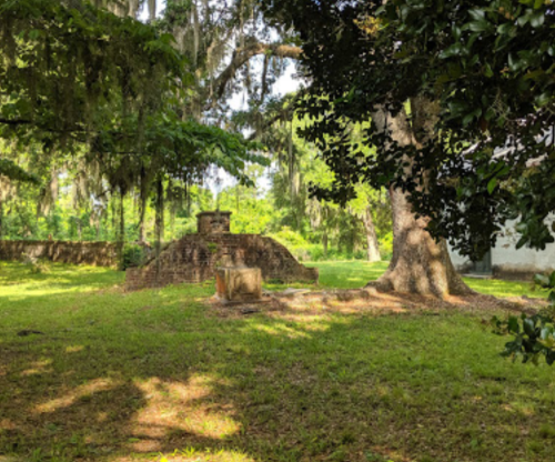 Ruins of a stone structure surrounded by lush greenery and trees, with dappled sunlight on the grass.