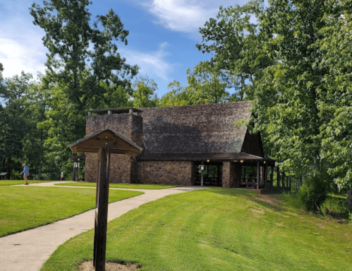 A stone building with a sloped roof surrounded by trees and a grassy area, with a path leading to it.