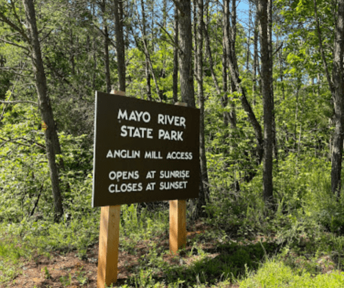 Sign for Mayo River State Park, Anglin Mill Access, with hours: opens at sunrise, closes at sunset, surrounded by trees.