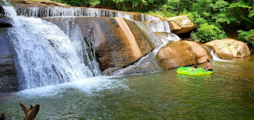 A serene waterfall cascades over rocks into a calm pool, with a person in a green kayak enjoying the water.