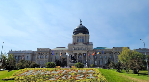 Historic building with a dome, surrounded by colorful flower beds and flags, under a clear blue sky.