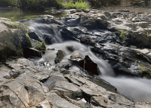 A serene stream flows over smooth rocks, surrounded by greenery in a tranquil natural setting.