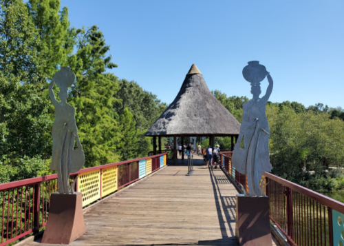 A wooden bridge leads to a thatched-roof pavilion, flanked by two sculptures of figures carrying pots.