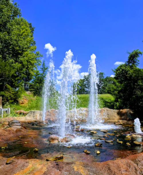 A vibrant fountain sprays water into the air, surrounded by greenery and a clear blue sky.