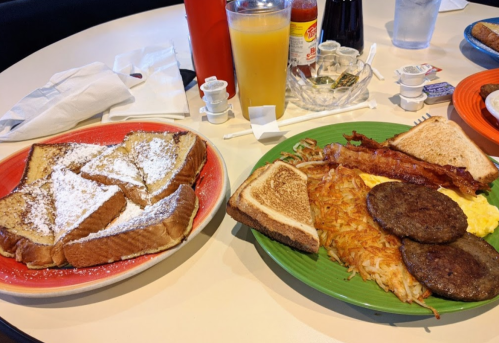 A table with two plates of breakfast: French toast with powdered sugar, and a plate with eggs, sausage, bacon, and toast.