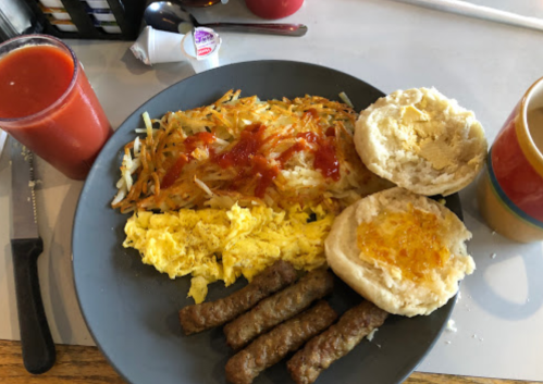 A plate of breakfast featuring scrambled eggs, hash browns with ketchup, sausage links, and a biscuit with jelly.