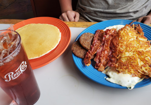A plate of breakfast with bacon, sausage, hash browns, and eggs, alongside a stack of pancakes and a glass of iced tea.