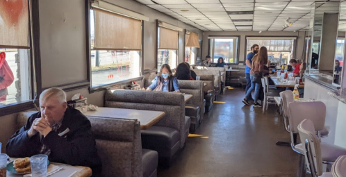 A diner interior with booths and tables, patrons eating, and a person in a mask sitting alone at a booth.