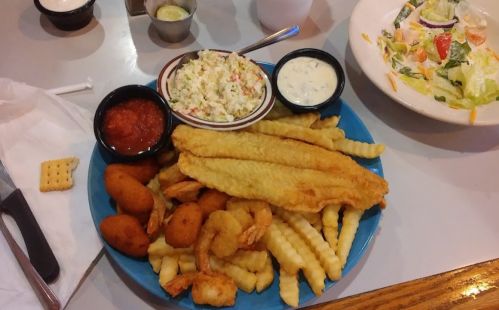 A blue plate filled with fried fish, shrimp, crinkle-cut fries, hushpuppies, coleslaw, and dipping sauces.