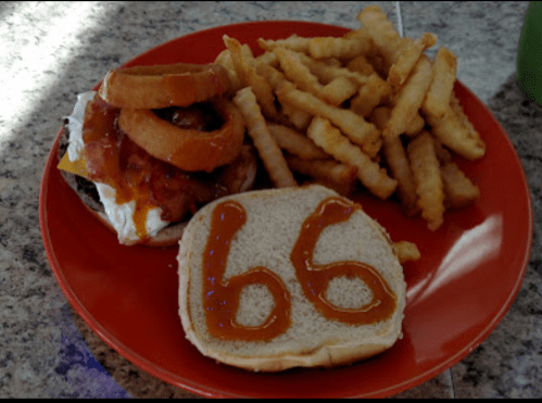A plate with a burger topped with onion rings and fries, alongside a slice of bread with the number "66" in ketchup.
