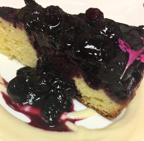 A slice of cake topped with a rich blueberry sauce and a small purple flower, served on a white plate.