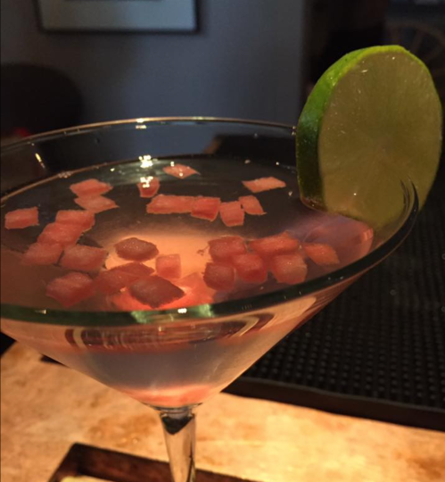 A martini glass filled with a clear drink, garnished with a lime slice and small pink cubes floating inside.