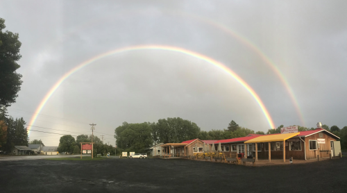A vibrant double rainbow arcs over a rustic building with a red roof, surrounded by greenery and a cloudy sky.