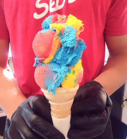 A person in a red shirt holds a colorful ice cream cone with blue, pink, and yellow scoops.