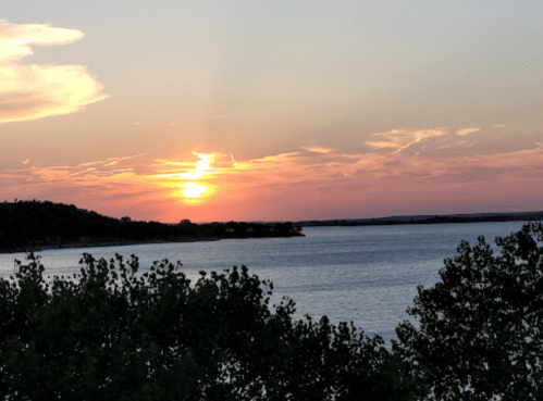 A serene sunset over a calm lake, with silhouettes of trees in the foreground and colorful clouds in the sky.