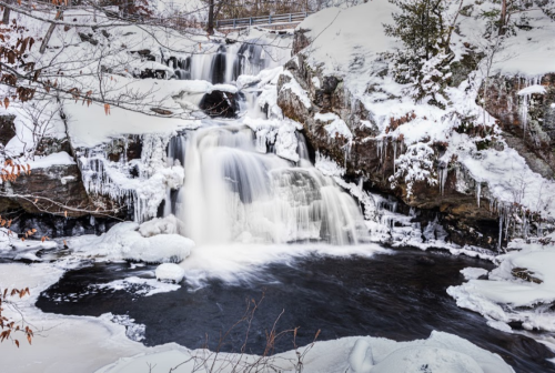 A frozen waterfall cascading over rocks, surrounded by snow and ice, with a dark pool at the base.