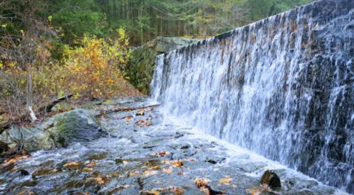 A serene waterfall cascading over a stone dam, surrounded by autumn foliage and a rocky riverbank.