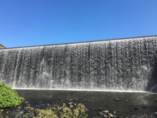A cascading waterfall flows over a dam under a clear blue sky, surrounded by greenery.
