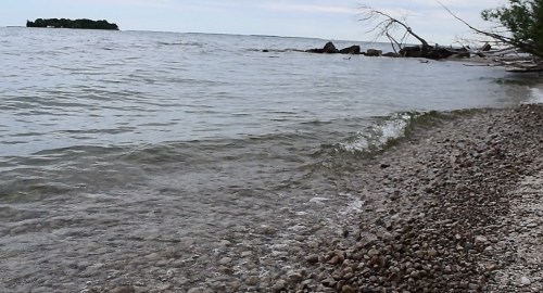 A calm shoreline with pebbles, gentle waves lapping at the water's edge, and a distant island on the horizon.