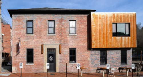 A modern brick building with a rust-colored metal addition, featuring large windows and outdoor seating.