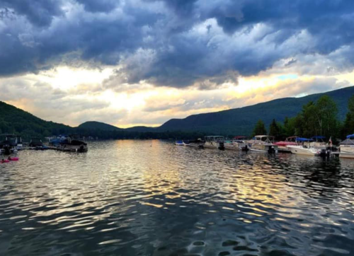 A serene lake at sunset, with boats docked and mountains in the background under a cloudy sky.