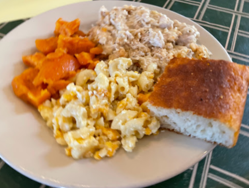 A plate with macaroni and cheese, shredded chicken, sweet potatoes, and a piece of cornbread on a green tablecloth.