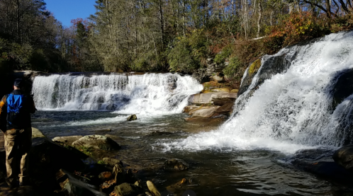 A person stands by a waterfall, surrounded by trees and rocks, with cascading water flowing over the rocks.