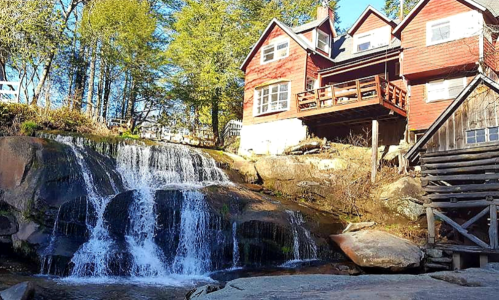 A charming red house beside a cascading waterfall, surrounded by lush green trees and rocky terrain.
