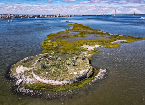 Aerial view of a small island with greenery, surrounded by water, with a bridge visible in the background.