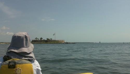 A person in a hat paddles a kayak towards a small island with a flag, surrounded by calm water and a clear sky.
