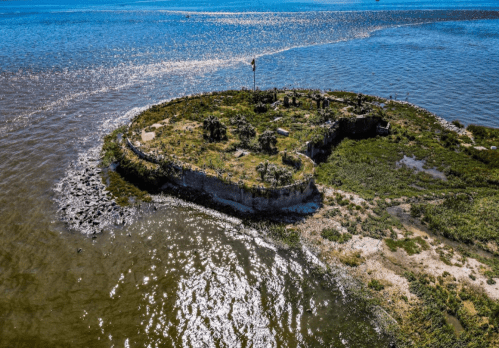 Aerial view of a small, grassy island surrounded by water, featuring a flag and rocky shoreline.