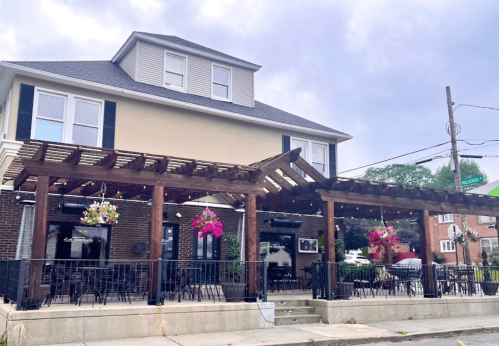 A charming restaurant with a wooden pergola, outdoor seating, and colorful hanging flowers, set against a cloudy sky.