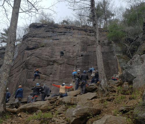 A group of climbers and instructors at a rocky climbing site, with one person ascending the cliff and others observing.
