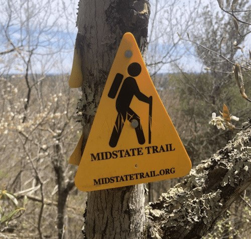 Yellow triangular sign for Midstate Trail, featuring a hiker icon and the website midstatetrail.org.